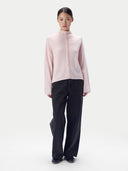A model stands against a light gray studio background wearing a pale pink mock-neck knit top with a decorative braided front detail and black wide-leg trousers.