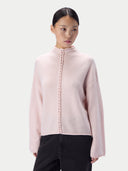 A model wearing a pale pink knit mock-neck sweater with a decorative braided front detail, paired with black pants, against a light gray studio background.