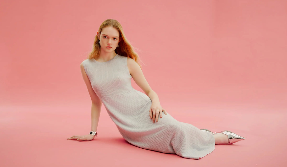 Frau in ärmellosem hellgrauem Feinstrick-Maxikleid und spitzen Silberschuhen auf rosa Boden, Frühling/Sommer 2024