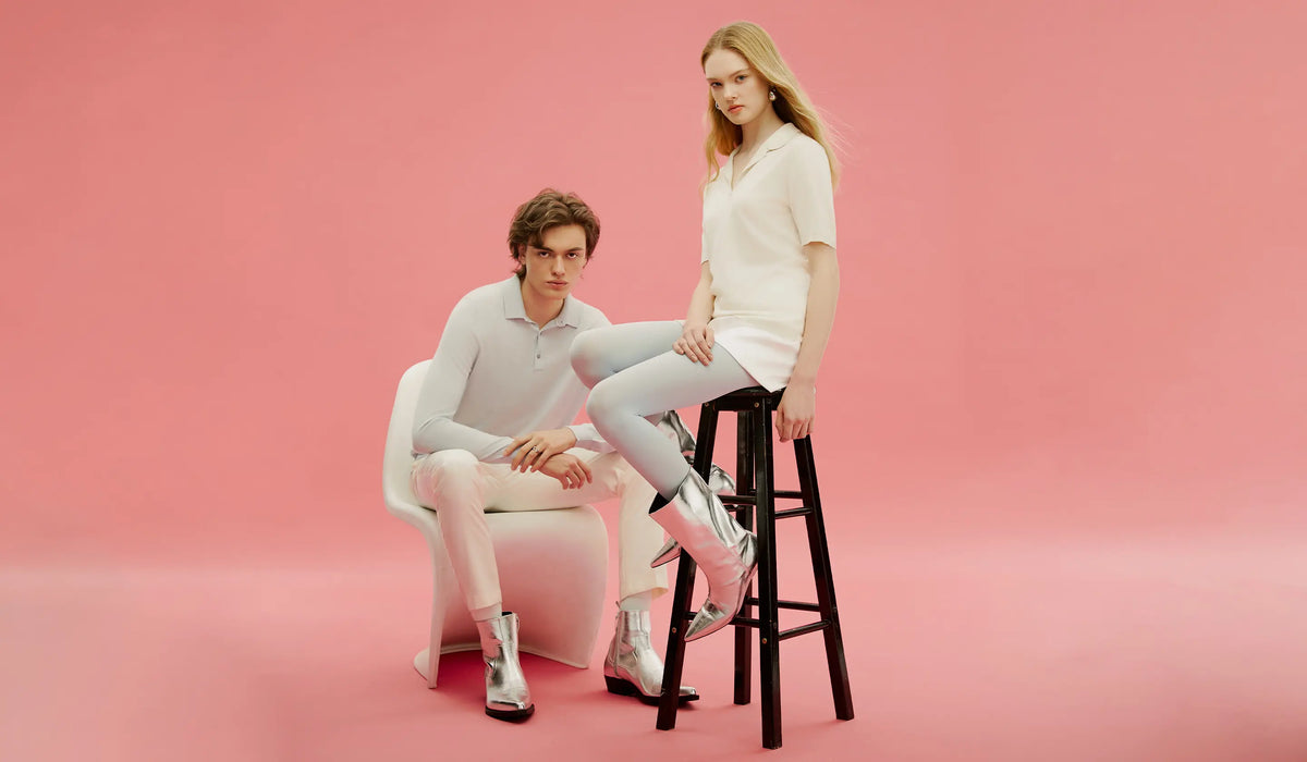 Mann und Frau in hellen Strick-Poloshirts und silbernen Stiefeln sitzen vor rosa Studiohintergrund, Frühjahr/Sommer 2024