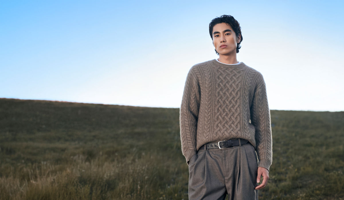 Mann in taupefarbenem Zopfstrick-Kaschmirpullover und grauer Hose auf einem Grashügel unter blauem Himmel