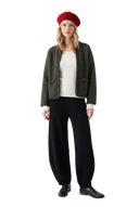 Frau in einer dunkelgrünen Kaschmir-Strickjacke mit handgeflochtenem Zopfmuster über einem weißen Rippstrick-Top, schwarze Kaschmir-Strickhose mit Tonnenbein, rote Strick-Beret und schwarze Loafer