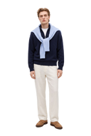 Mann in einem dunkelblauen Kaschmir-Troyer mit Reißverschluss, hellblauer Zopfstrick-Pullover über den Schultern, cremefarbene Hose und hellbraune Loafer