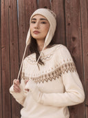 Frau in cremefarbenem Fair-Isle-Kaschmirpullover mit passender Ohrenklappenmütze vor einer roten Holzwand