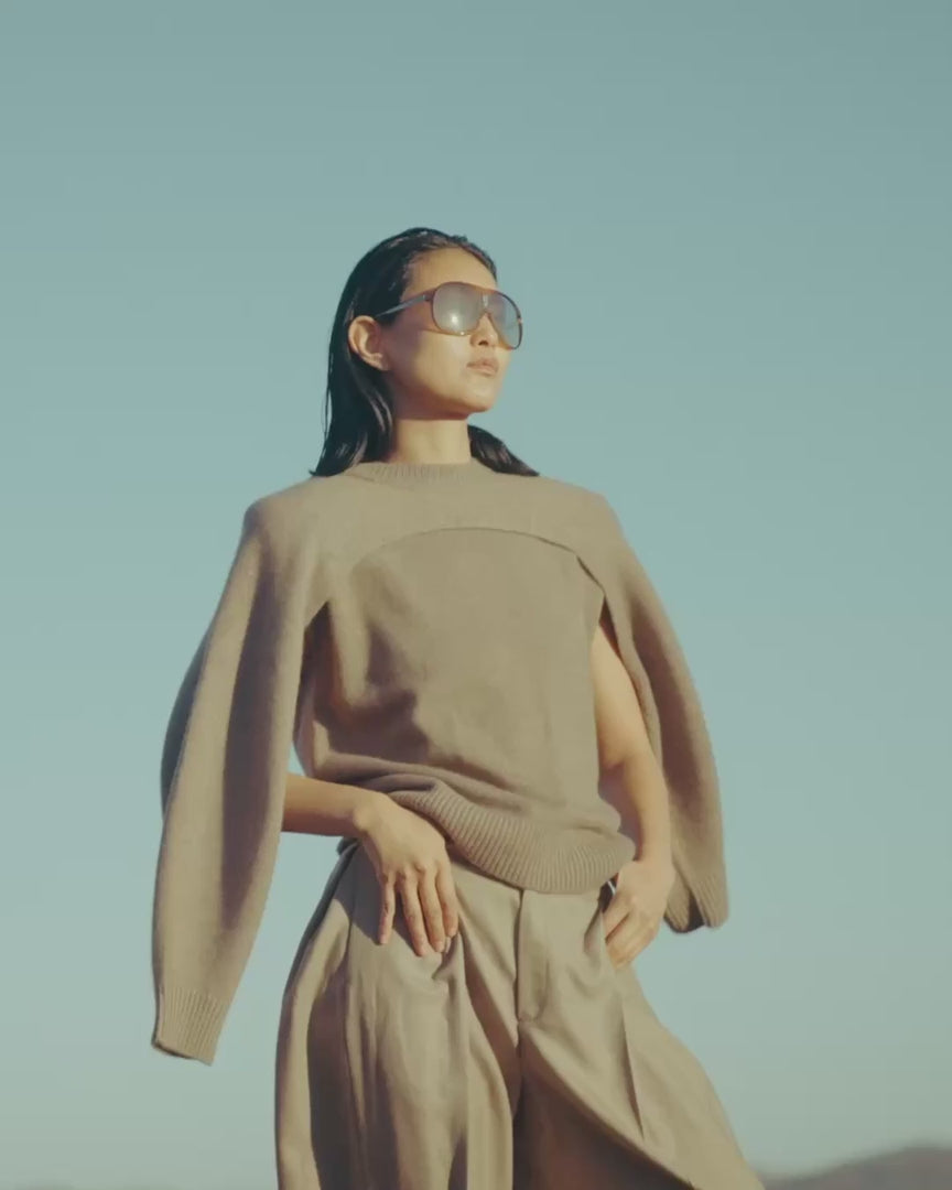 Frau mit Sonnenbrille in taupefarbenem Kaschmirstrick mit drapiertem Bolero und weiter Hose vor klarem blauem Himmel